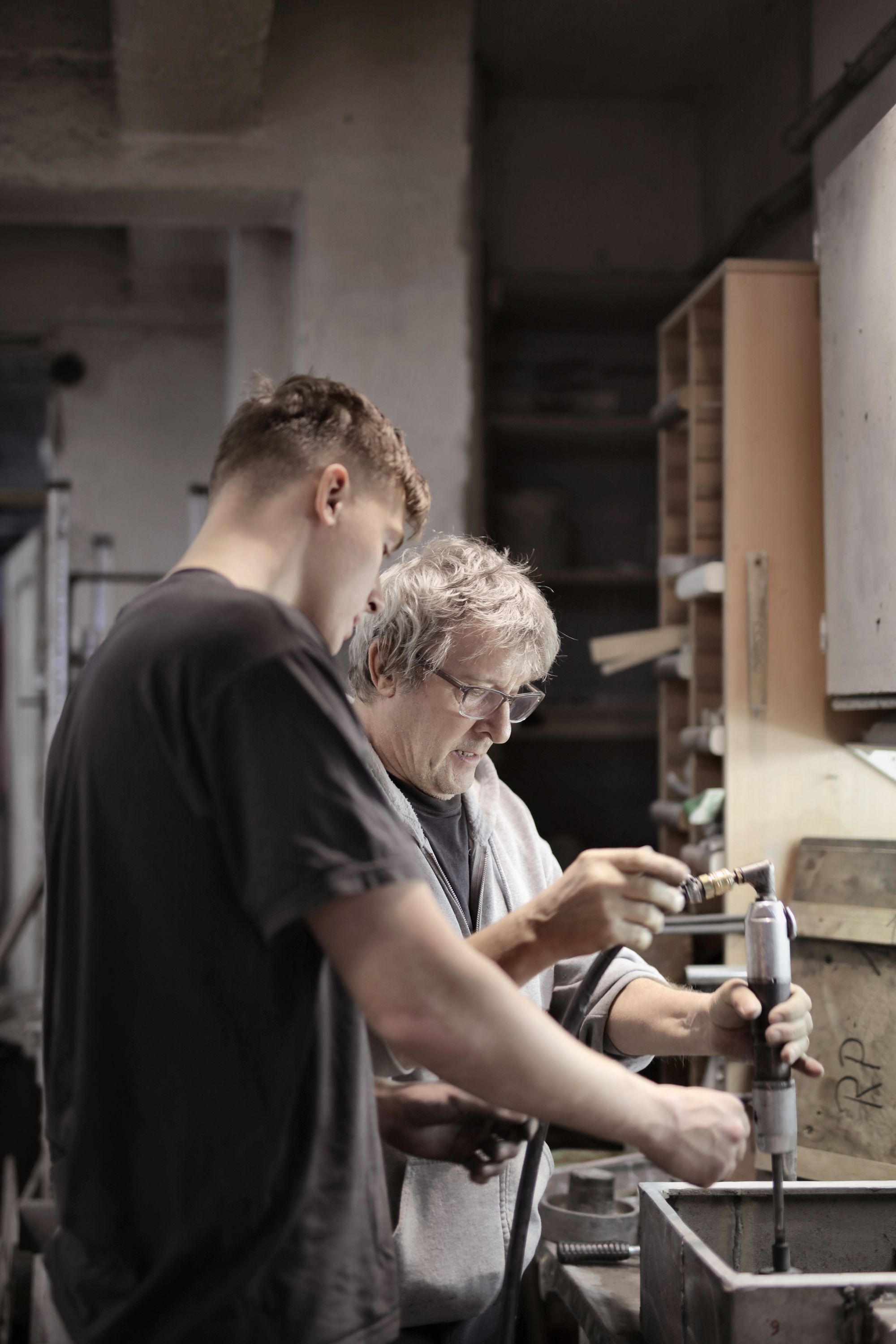 Erfahrener Werkstattmeister mit Brille setzt in einer Werkstatt ein Druckluftwerkzeug an, während sich ein junger Auszubildender über seine Schulter beugt und aufmerksam zuschaut, Symbolbild für Teambelege auf einer Karriereseite