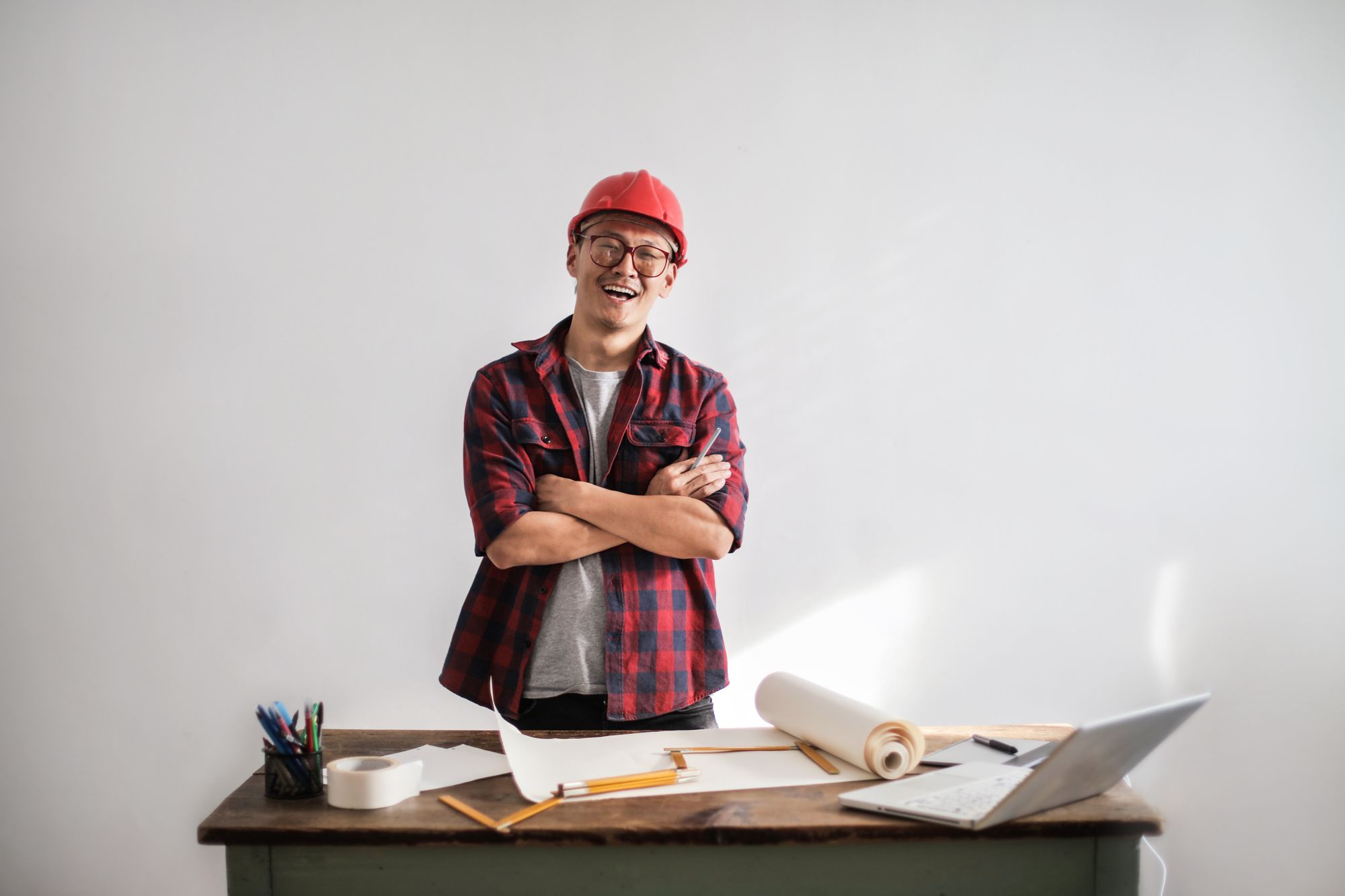 Lachender Handwerker mit rotem Schutzhelm, Brille und kariertem Flanellhemd steht mit verschränkten Armen an einem Holztisch mit Bauplänen, Stiften, Linealen und Laptop vor heller Wand, Symbolbild für die planende Verantwortung für den Recruiting-Prozess