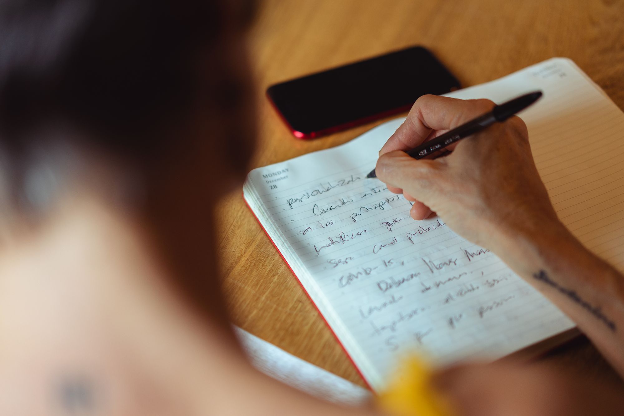 Hand schreibt mit schwarzem Stift in ein liniertes Notizbuch neben einem Smartphone auf einer Holzfläche