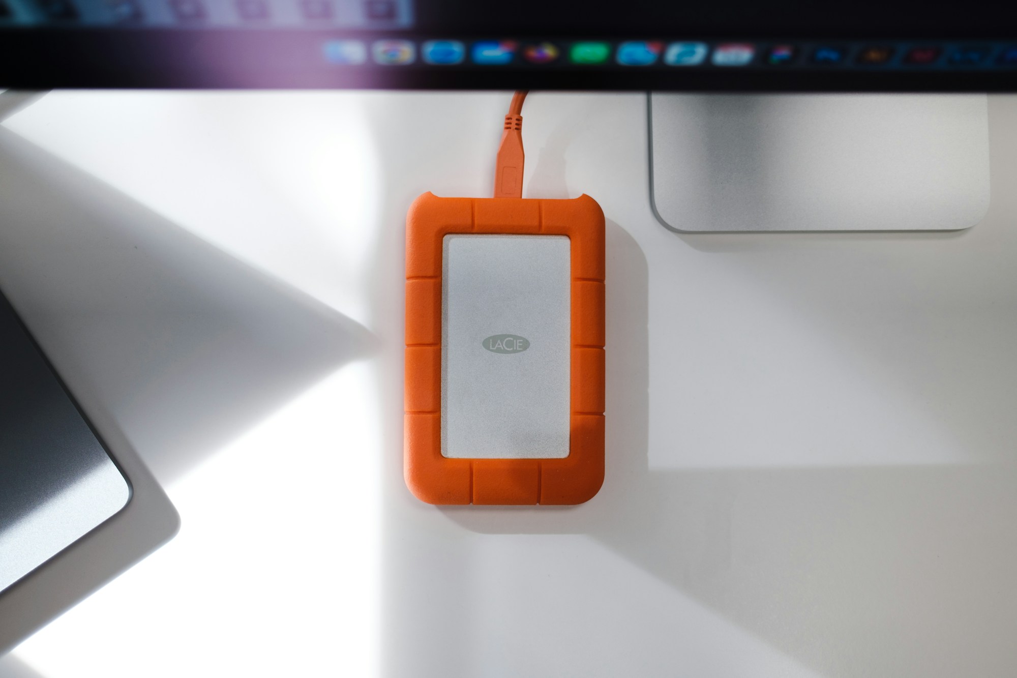Externe Festplatte mit orangefarbenem Schutzgehäuse und Kabel auf einer weißen Fläche vor einem Bildschirm